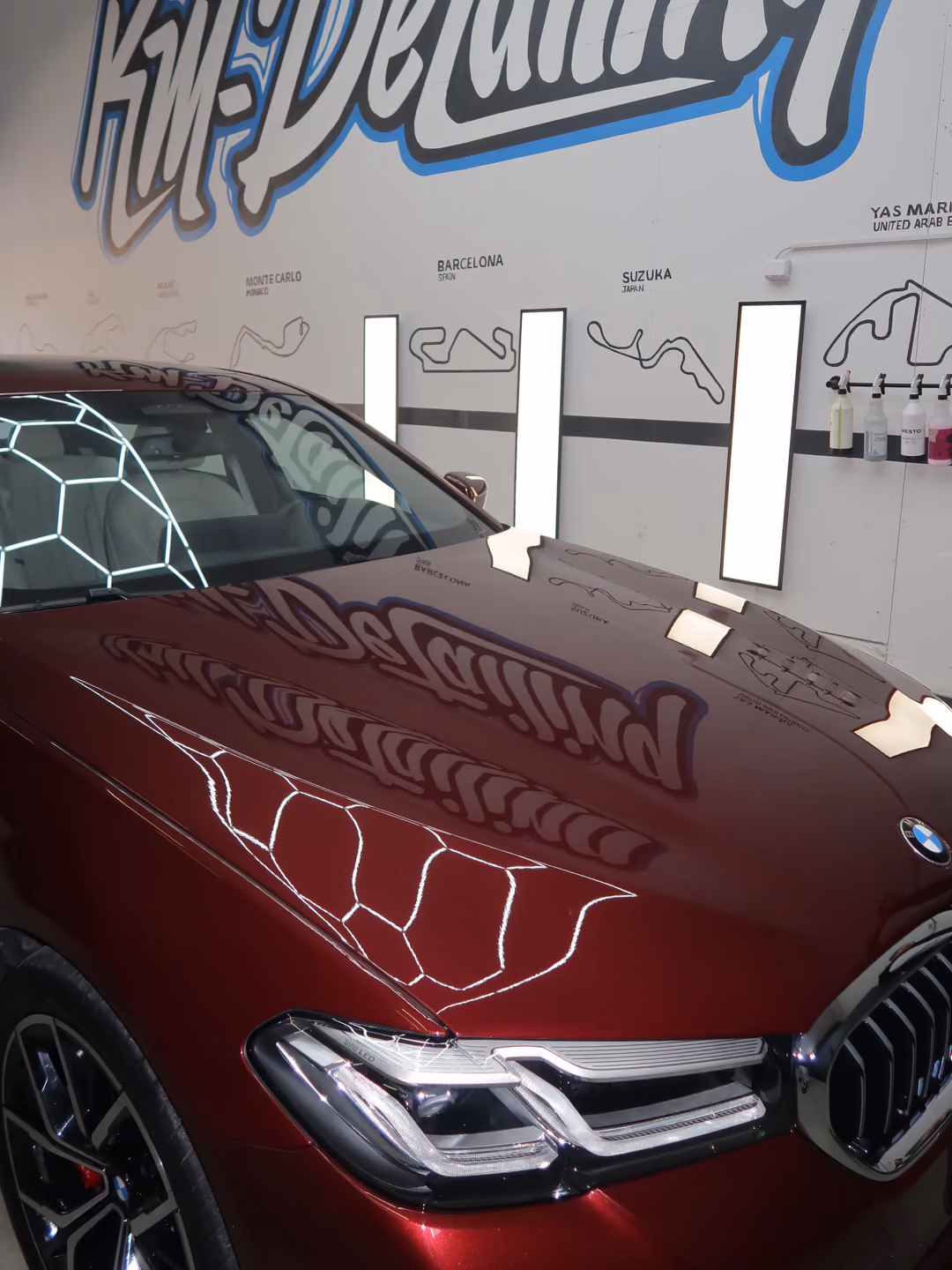 Viininpunaisen BMW 5-sarjan konepelti pinnoituksen jälkeen: peilikirkkaasta maalipinnasta heijastuvat KM-Detailingin logo ja kuusikulmiovalot